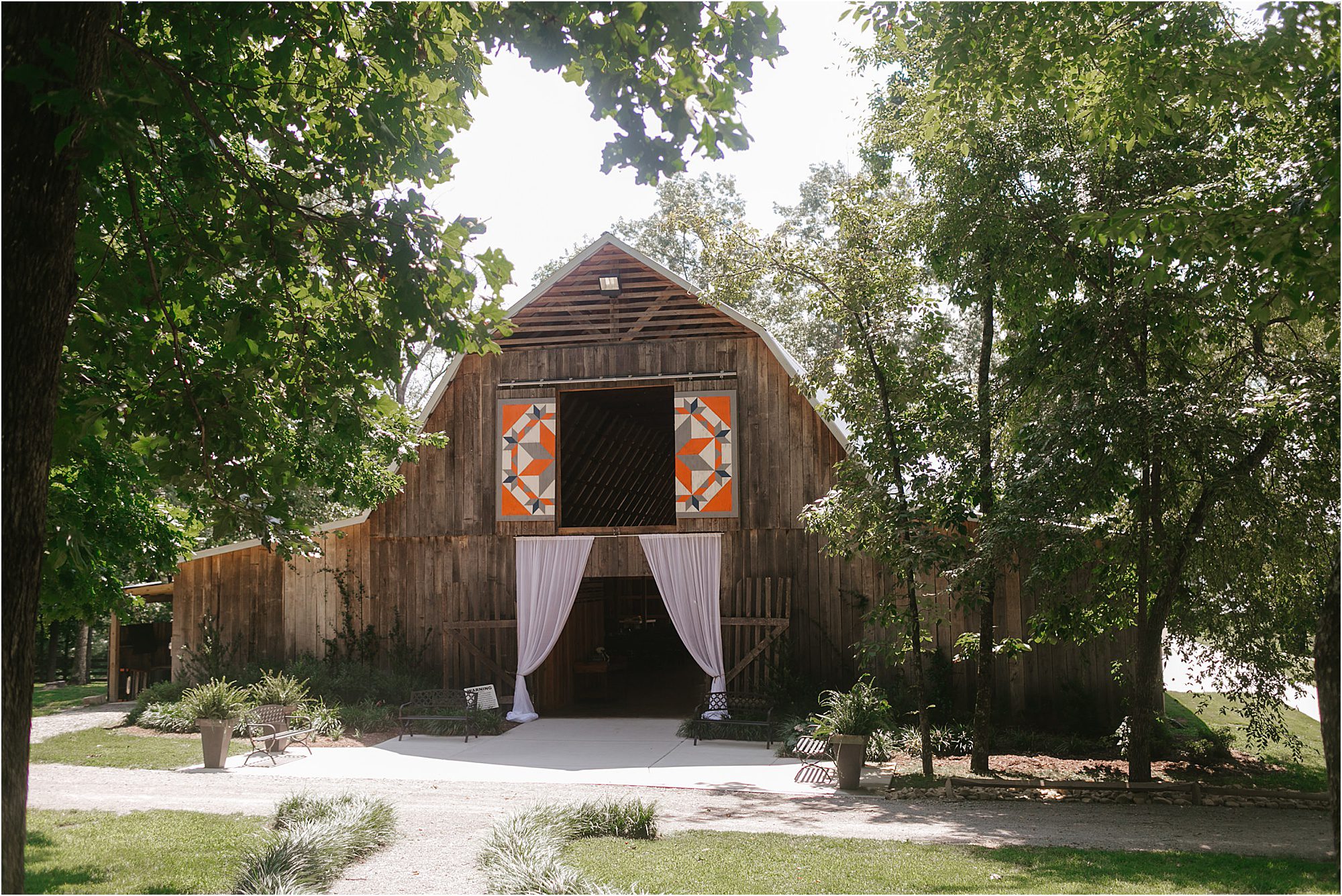 A rustic wooden barn at Riverview Family Farm features orange and white quilt art above the entrance, draped white curtains, and is surrounded by green trees and plants, with a gravel path in front.