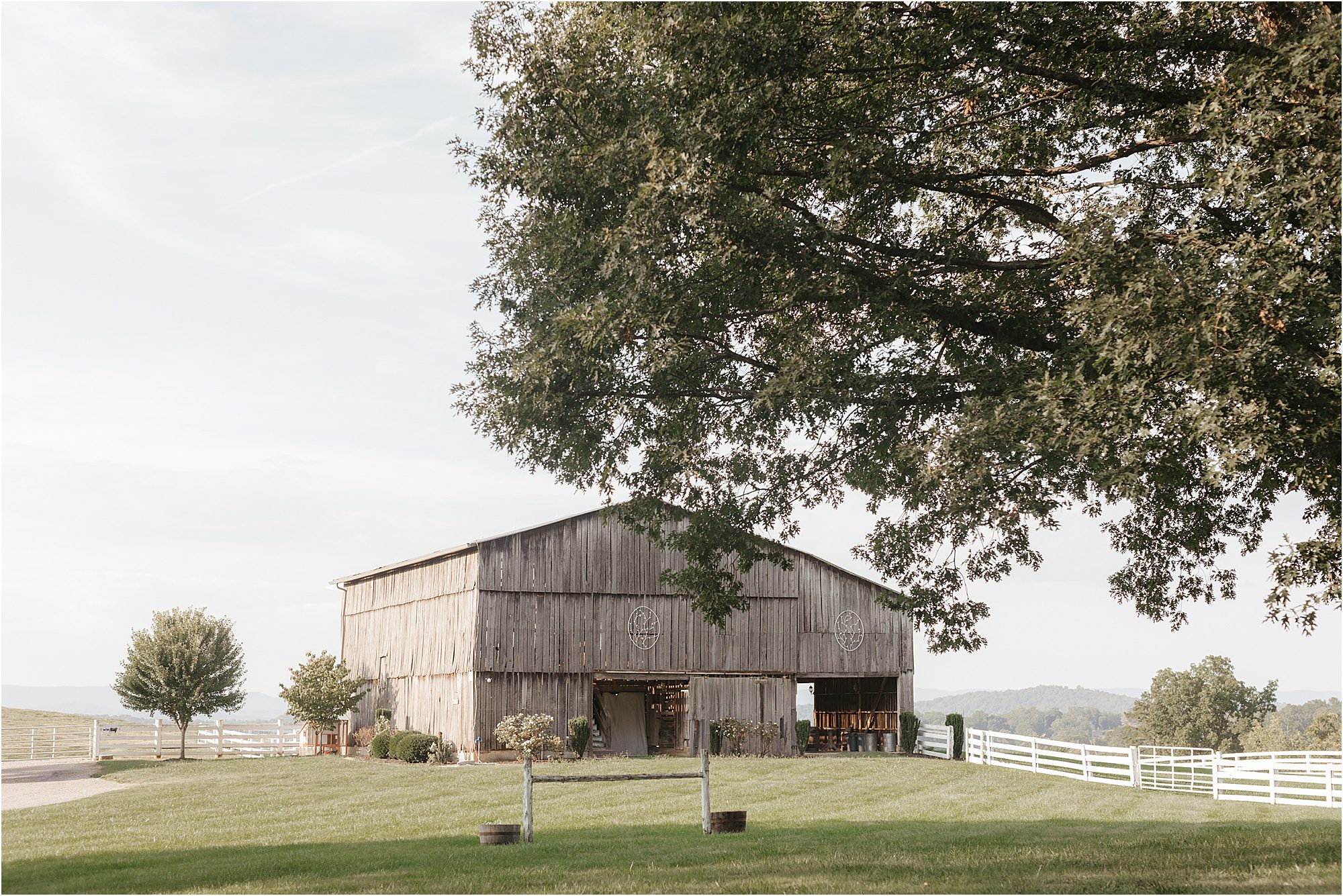 A rustic wooden barn stands in a grassy field at Hidden Meadows Farm, partially shaded by a large tree. White fences and distant hills are visible under a bright, clear sky.