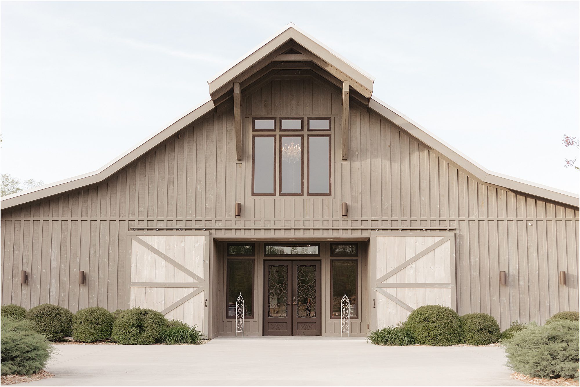 A large, rustic barn with wood siding, double glass doors, and tall windows stands proudly at Howe Farms. The barn is flanked by green bushes and has symmetrical sliding doors on each side.