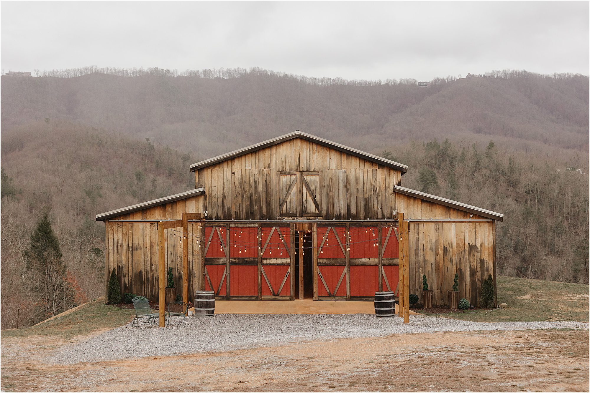 A rustic wooden barn with large red doors stands in front of rolling, tree-covered hills under an overcast sky at Mountain Mist Farms. Gravel and sparse grass cover the ground.
