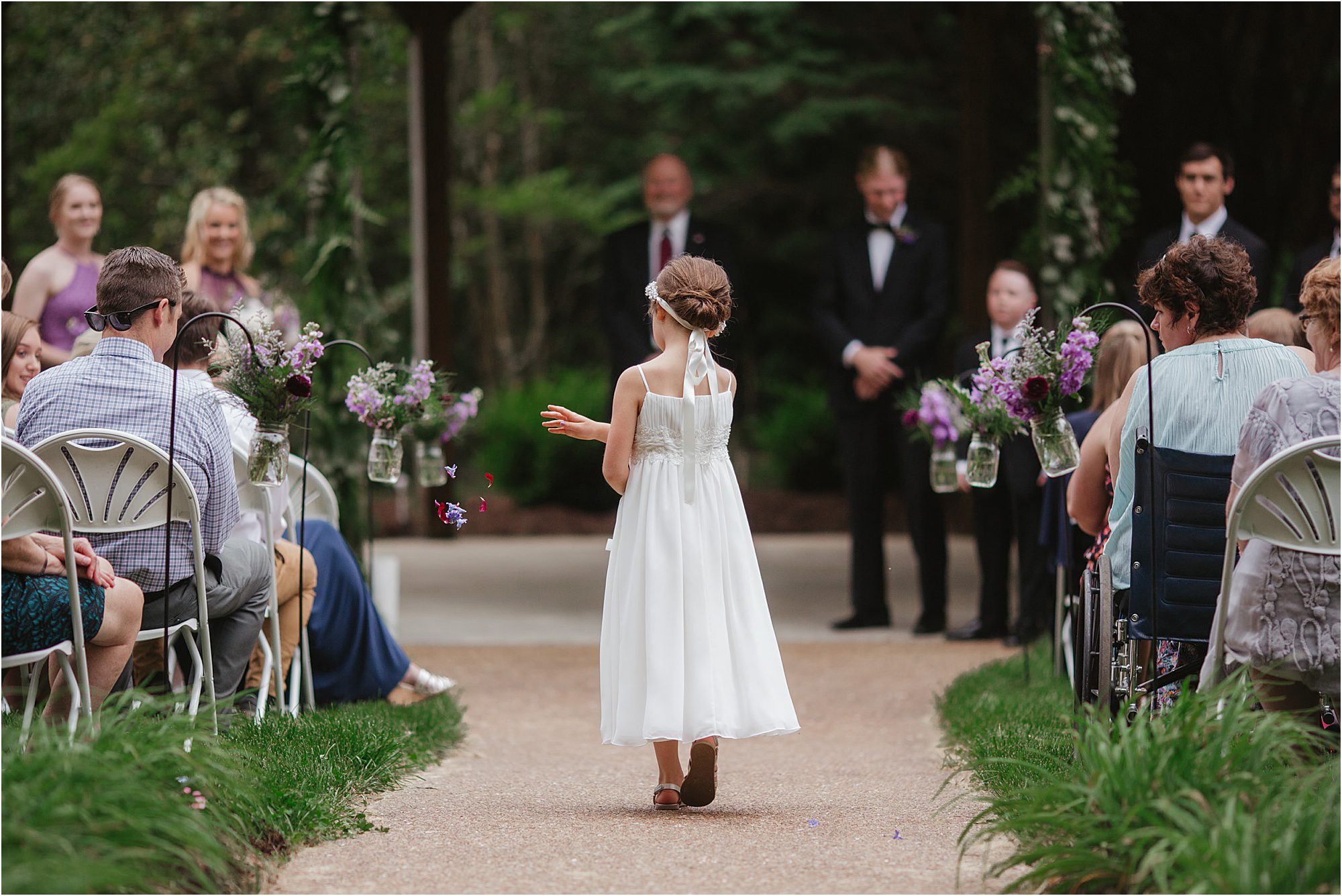 A young flower girl in a white dress walks down the outdoor wedding aisle at the Lily Barn, scattering petals, while guests sit on either side and the wedding party waits at the altar ahead.