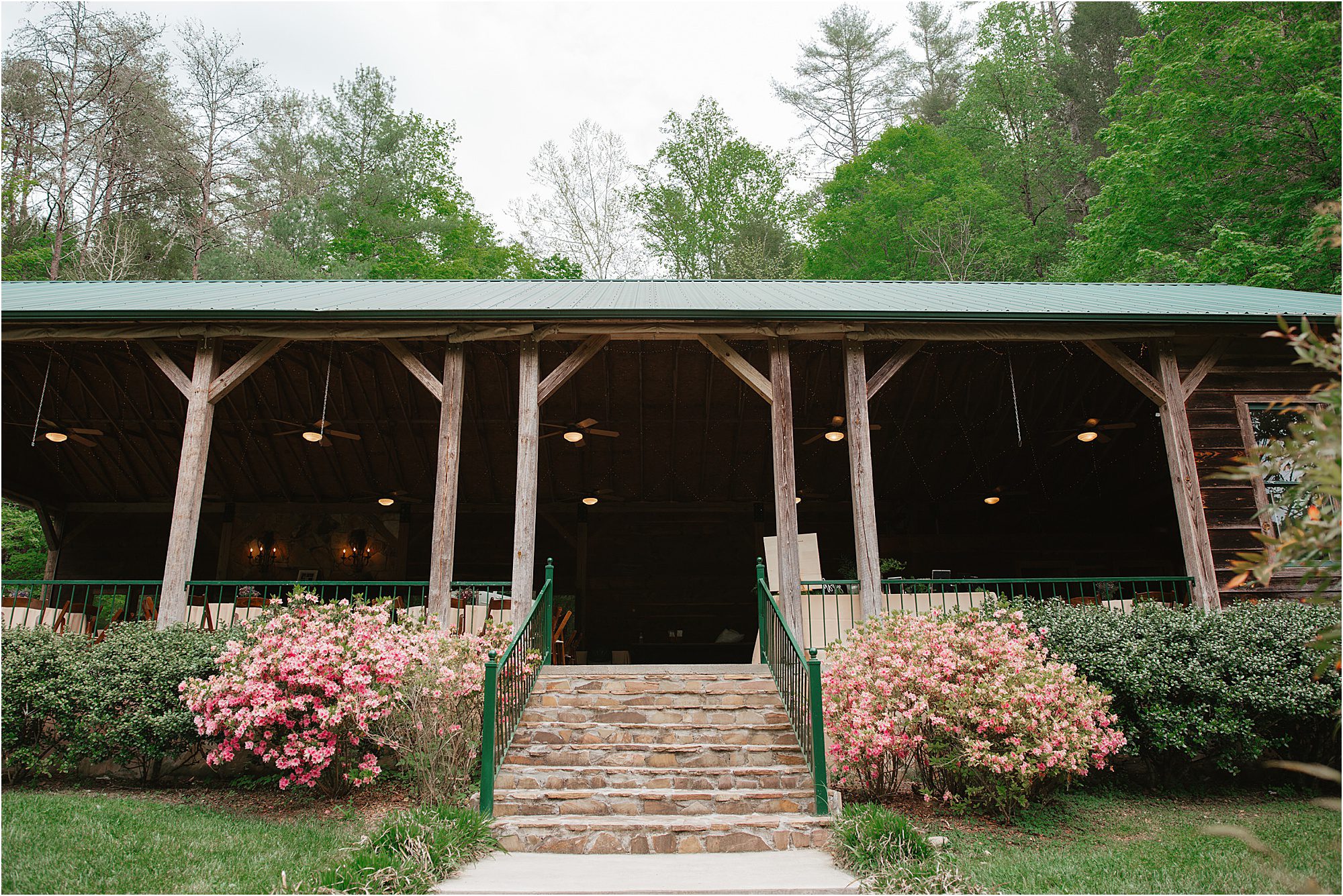 A rustic wooden pavilion with a green metal roof sits behind stone steps at the Lily Barn, surrounded by blooming pink and white bushes and lush green trees.