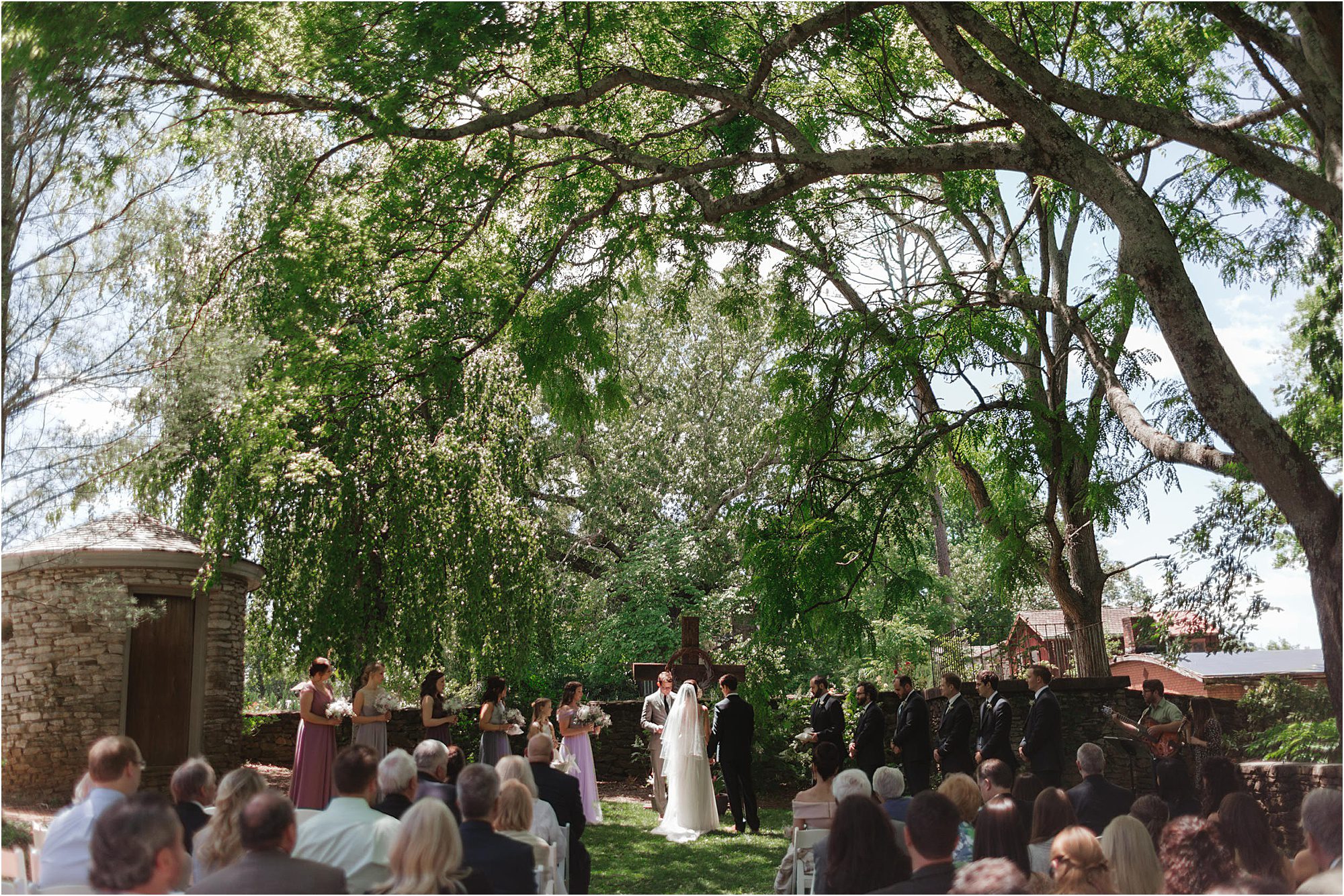 A wedding ceremony takes place outdoors under large trees at the Knoxville Botanical Garden and Arboretum, with the couple at the altar, bridesmaids and groomsmen standing on either side, and guests seated in rows. Sunlight filters through the leaves.