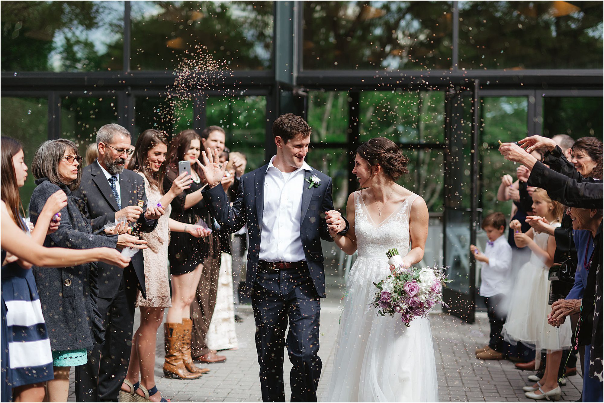 A newlywed couple smiles and holds hands as guests throw confetti outside at the Knoxville Botanical Garden and Arboretum, celebrating their wedding. The bride holds a bouquet and wears a white dress; the groom is in a suit. People cheer on both sides.