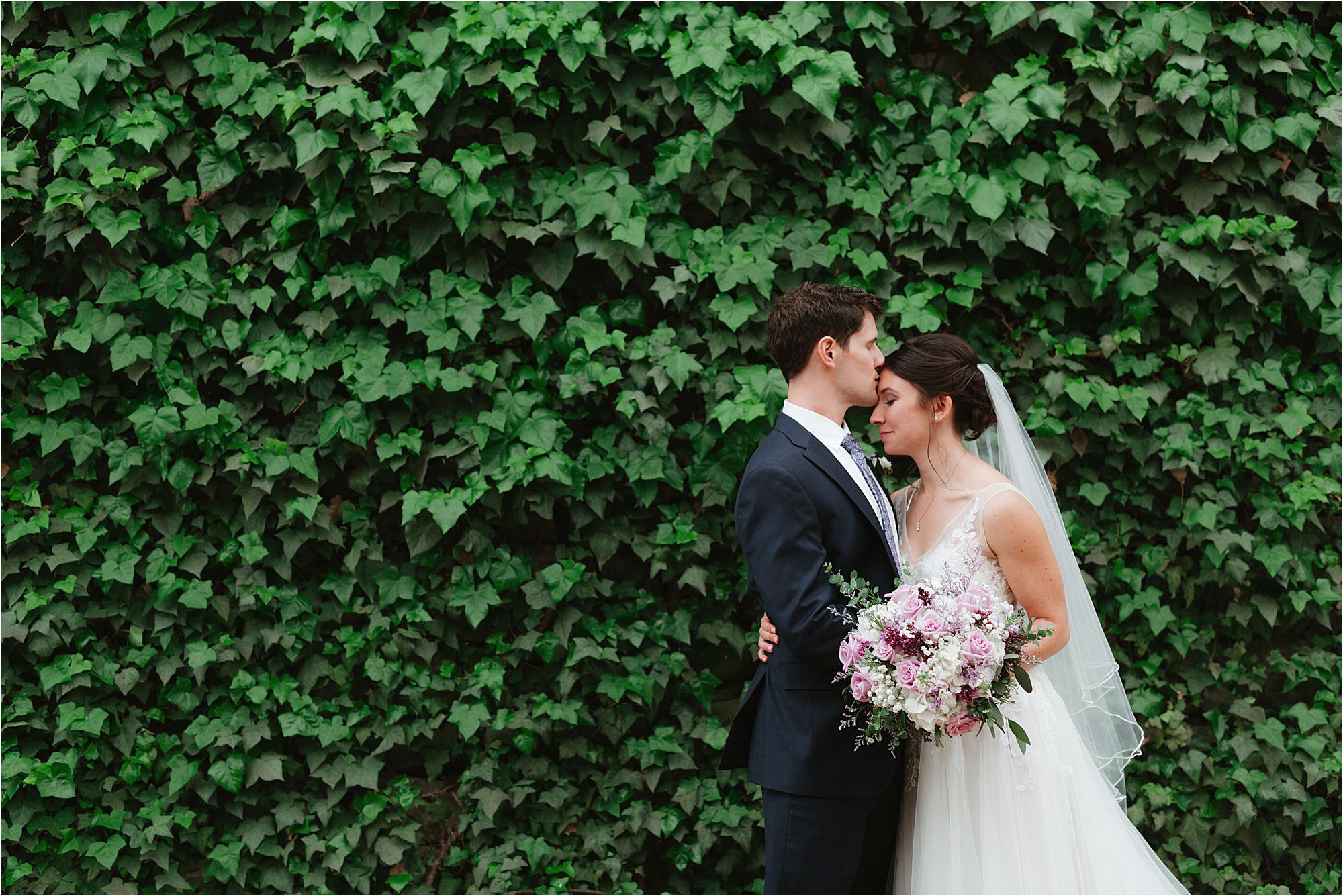 A bride and groom embrace in front of a lush green ivy wall. The bride holds a bouquet of pink and white flowers and wears a white veil, while the groom wears a dark suit and tie.