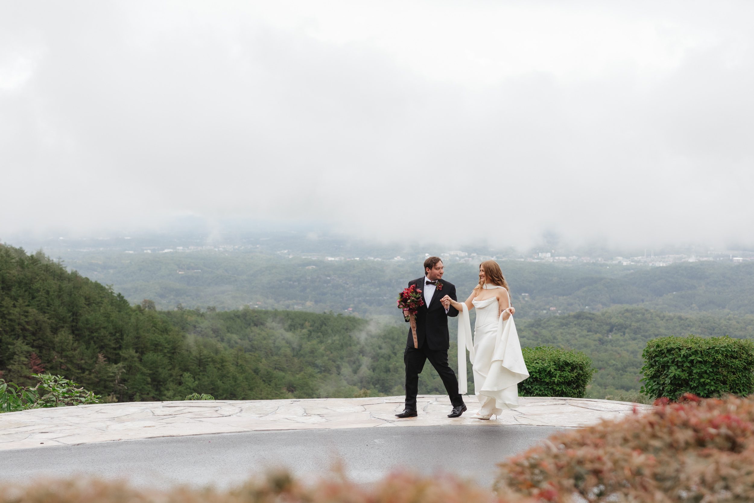 A bride and groom in wedding attire walk hand-in-hand on a stone patio at The Trillium Venue, overlooking a misty, mountainous landscape.