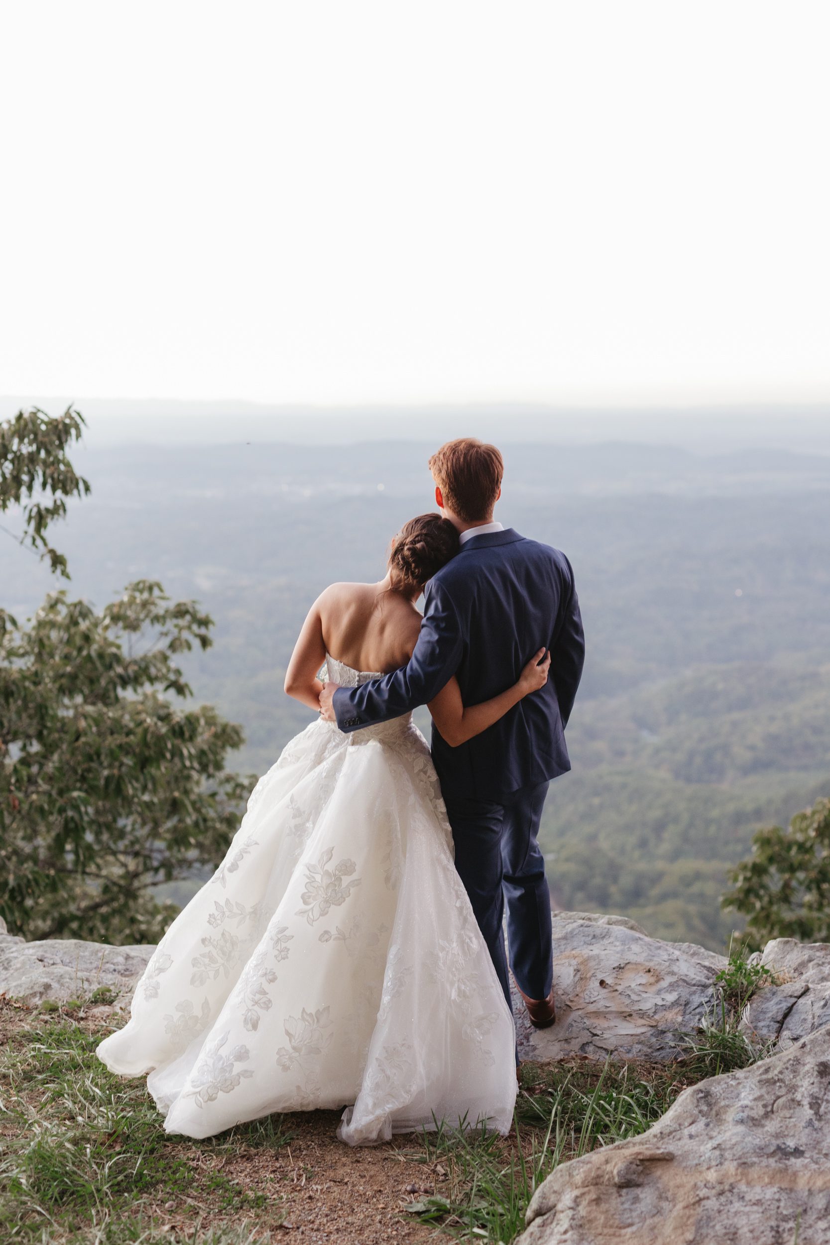 A bride in a white wedding gown and a groom in a navy suit stand on a rocky overlook, looking out over a mountain valley.